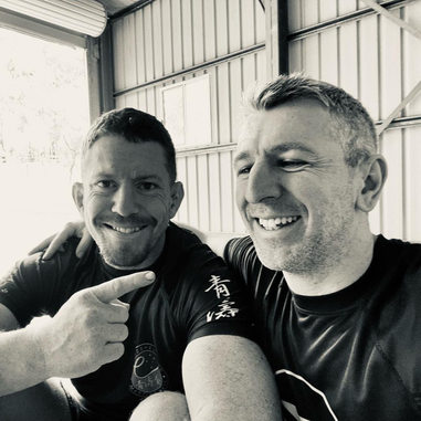 Two men sitting together in a gym, smiling as one points at the other in a friendly moment.