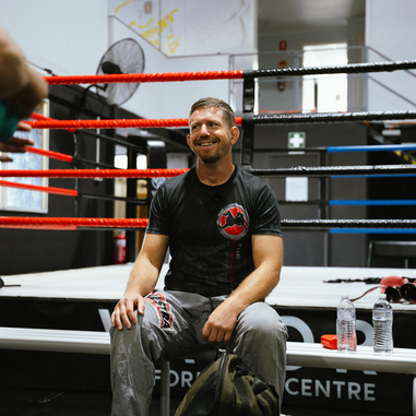 A man sitting on a bench beside a boxing ring, smiling and relaxing after training.