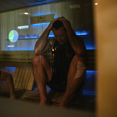 A man sitting in a sauna with his elbows on his knees and hands on his head, sweating during a heat therapy session.