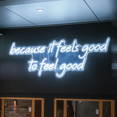 A glowing neon sign reading “because it feels good to feel good” mounted on a dark interior wall.