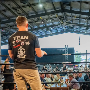 A man standing inside a boxing ring addressing a seated audience at a charity boxing event.